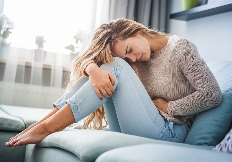 übersäuerung kann zu schmerzen führen, blonde frau krümmt sich auf dem sofa mit schmerzen