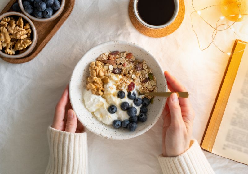 Hände, die eine Schüssel mit Joghurt, Müsli und Blaubeeren halten